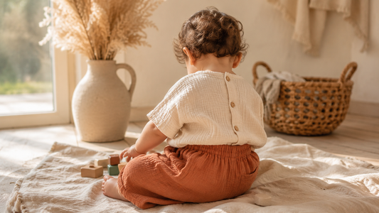 Bébé vu de dos portant un pantalon sarouel terracotta et un haut en gaze de coton crème dans une chambre bohème lumineuse.