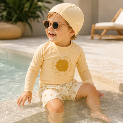 Bébé portant un ensemble de bain jaune avec manches longues, short à motifs et bonnet assorti, assis au bord d’une piscine avec des lunettes de soleil.