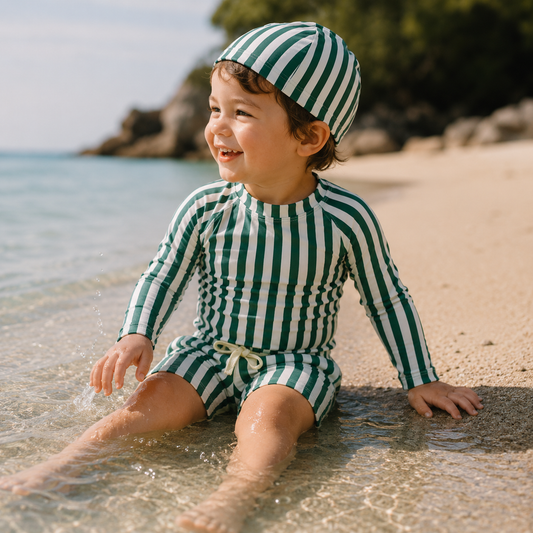 Enfant portant un maillot de bain rayé vert à manches longues avec bonnet assorti, assis au bord de l’eau sur la plage.