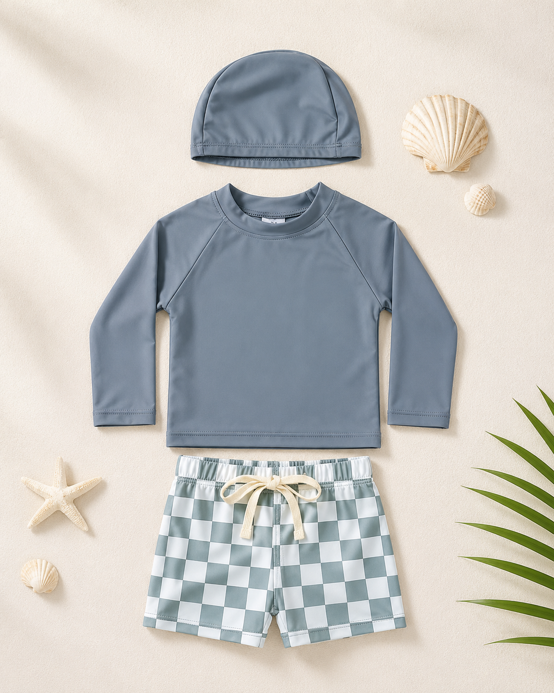 Ensemble de bain bébé gris à manches longues avec short à carreaux et bonnet assorti, présenté à plat dans une mise en scène plage minimaliste.