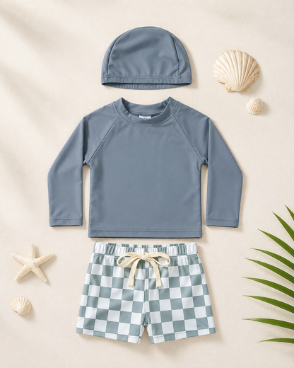 Ensemble de bain bébé gris à manches longues avec short à carreaux et bonnet assorti, présenté à plat dans une mise en scène plage minimaliste.
