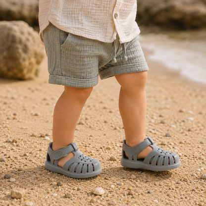 Sandales gladiateur bébé grises portées par un petit garçon sur une plage au style bohème naturel.