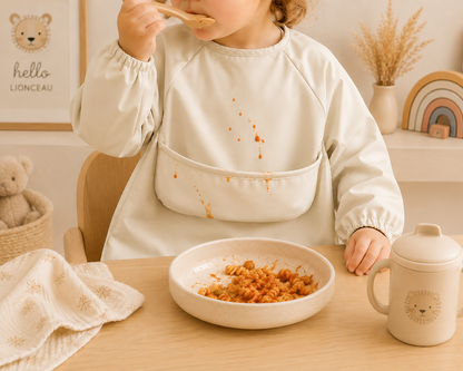 Enfant portant un tablier de peinture imperméable beige sable pendant un repas, avec éclaboussures de sauce sur le devant dans une ambiance bohème chaleureuse.