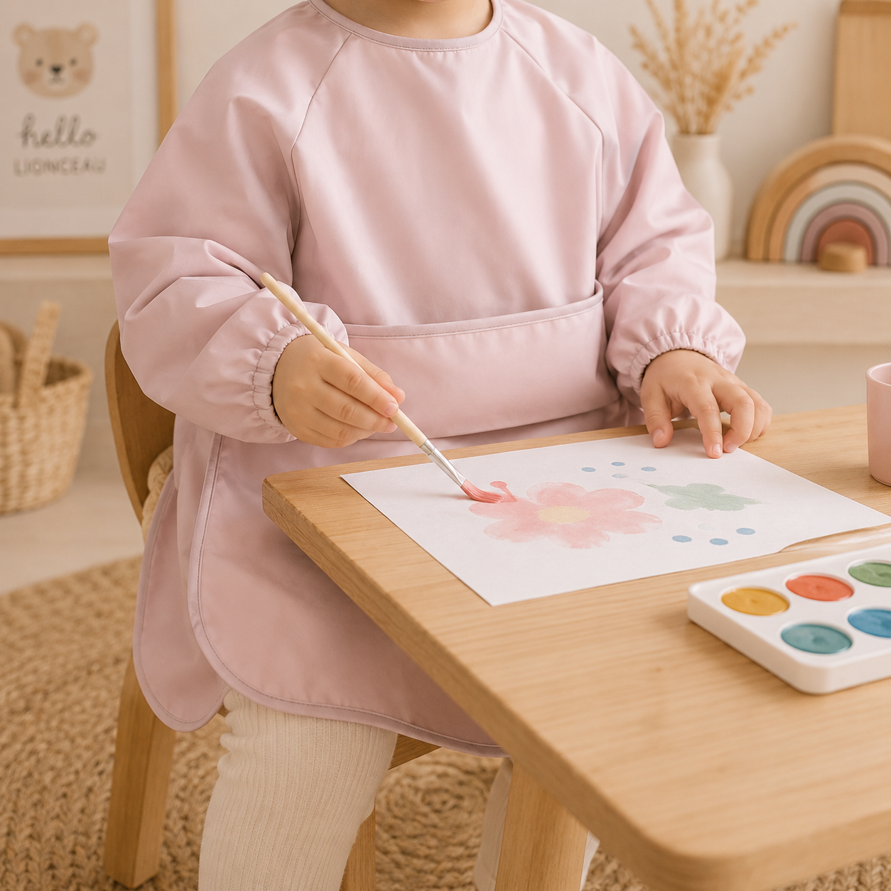 Enfant portant un tablier de peinture rose poudré en train de peindre à une table en bois dans une chambre bohème lumineuse.