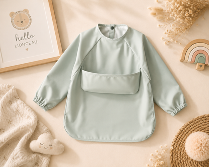 Tablier imperméable vert sauge pour bébé et enfant présenté dans une mise en scène bohème aux tons beige et naturels avec décoration Hello Lionceau.